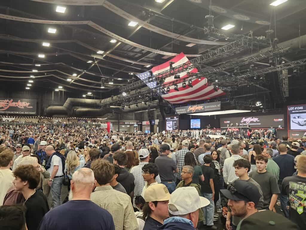 Large crowd gathers at a classic car auction with a stage and American flag backdrop.
